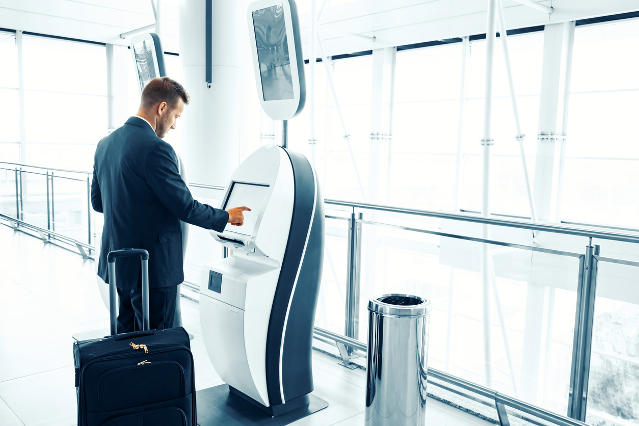 Business traveler using a self-service kiosk at an airport, processing airline compensation claim, with a suitcase beside him.